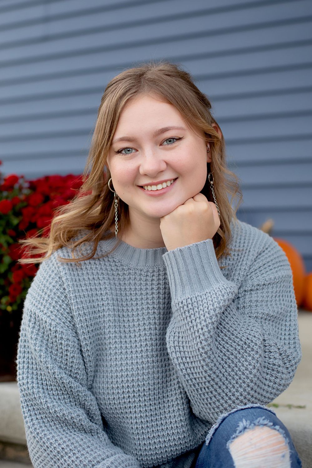 Outdoor portrait with red mums and blue siding, wearing a chunky grey sweater and dangle earrings.