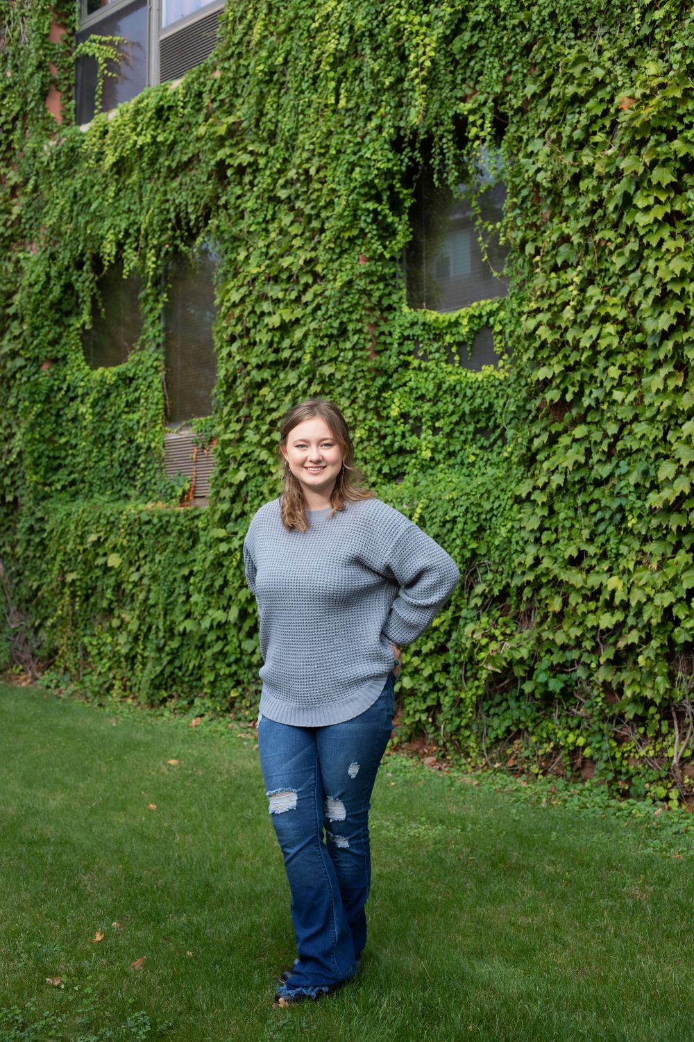 Standing on grass with ivy-covered building backdrop in casual grey sweater and jeans.