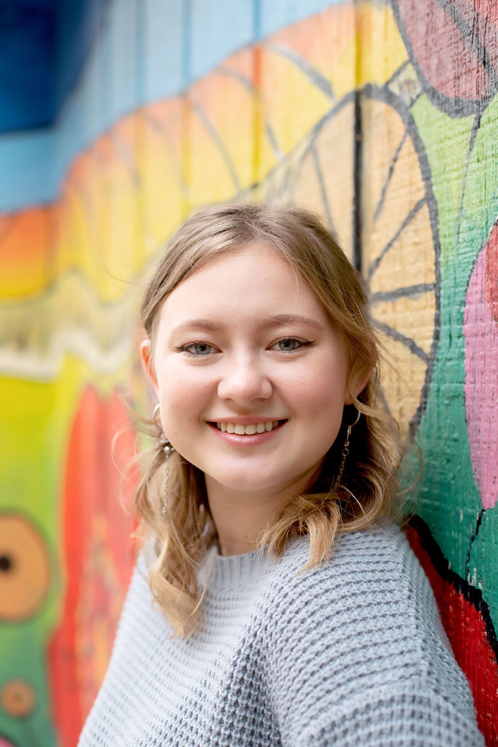 Person wearing gray sweater smiles in front of bright floral street art.