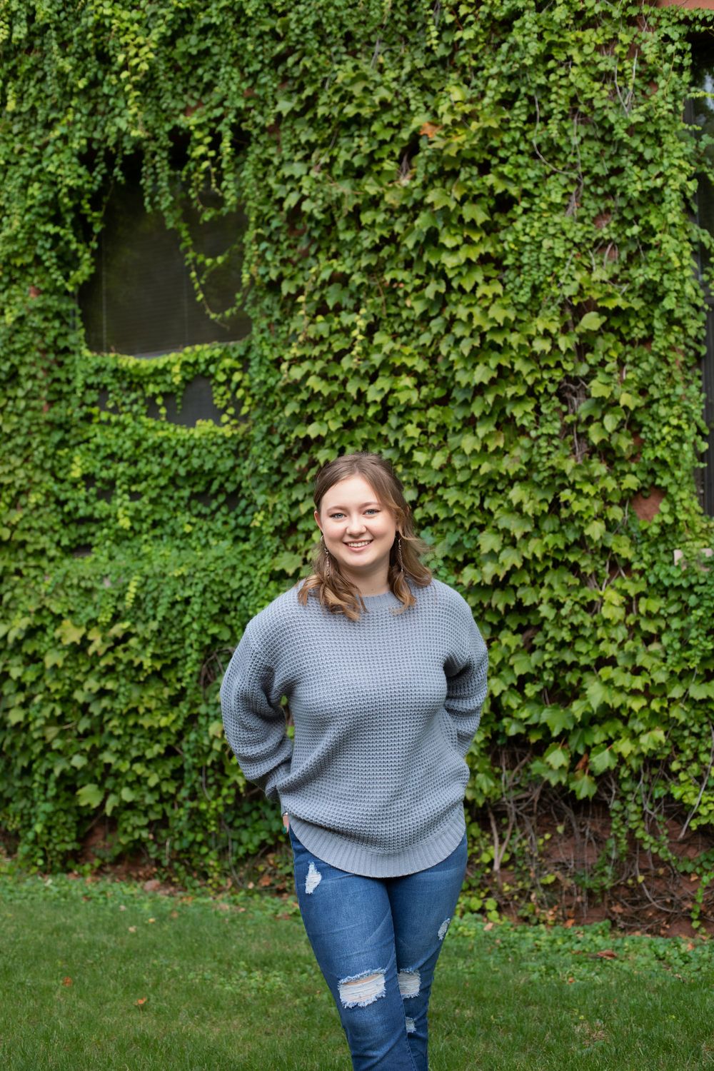 Standing in front of an ivy-covered wall wearing a grey sweater and distressed jeans.