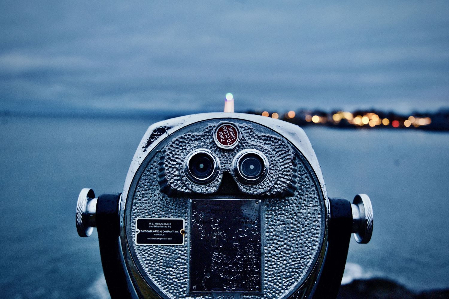 Coin-operated binocular viewer overlooking a coastal city at dusk with lights twinkling in the distance.