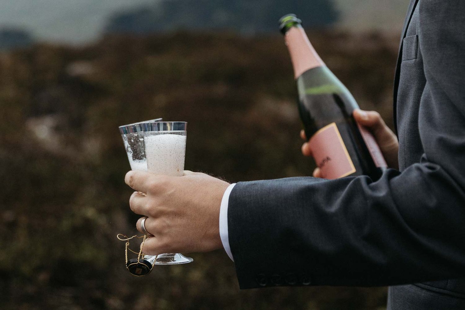 Hand in suit holding champagne bottle and glass against blurred landscape.
