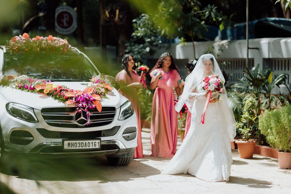 Wedding party poses outdoors with luxury car and floral arrangements in garden setting.