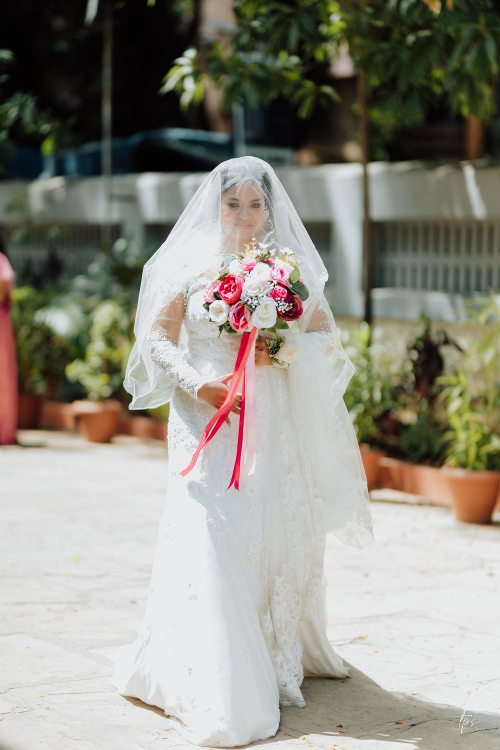 Bride in white lace gown holds pink and red bouquet in outdoor garden setting.