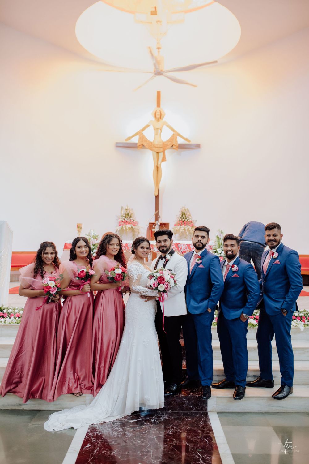 Wedding party poses in navy blue suits and dusty rose bridesmaid dresses in an elegant white venue.