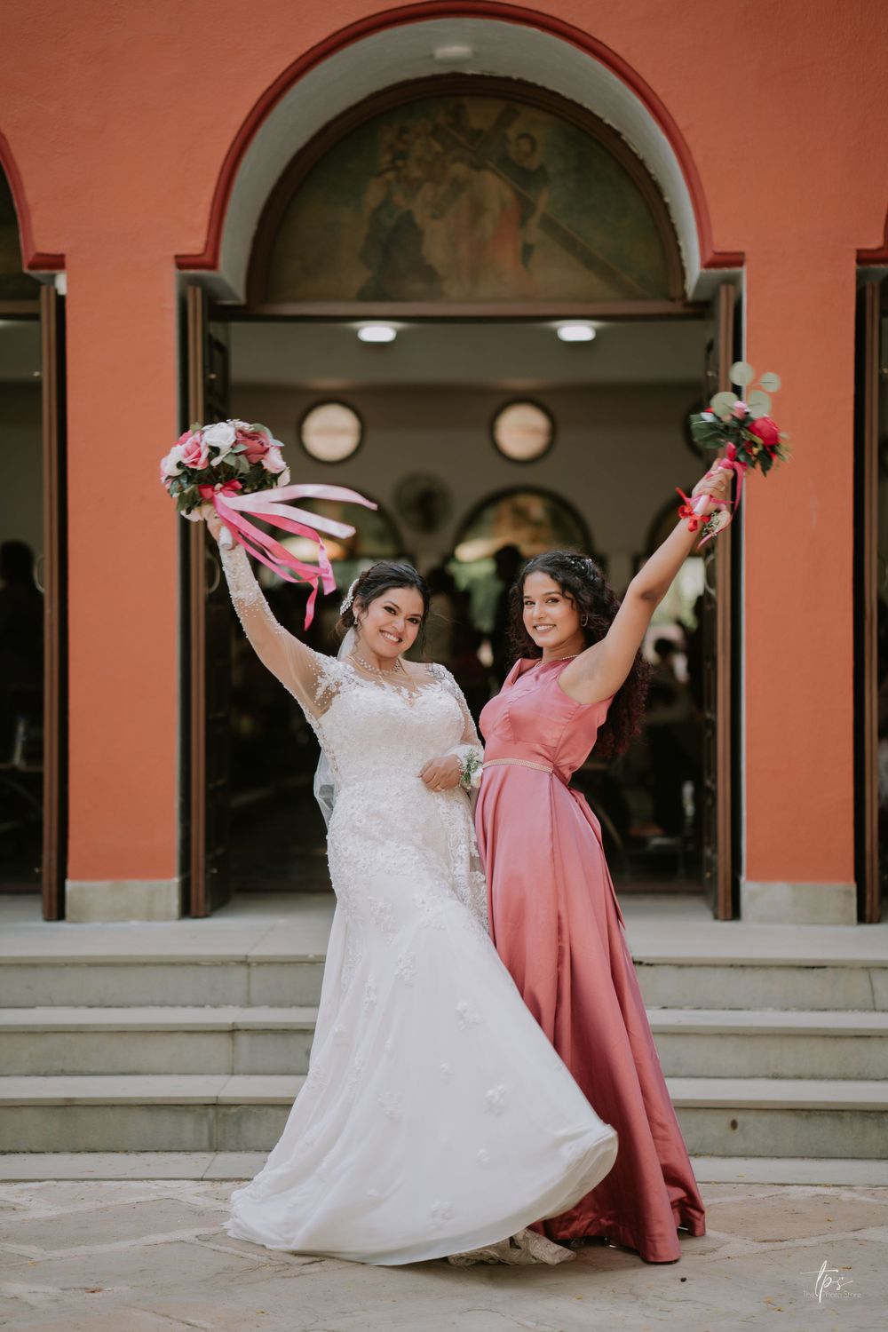 Bride and bridesmaid in dusty rose dress dance together under coral arches.