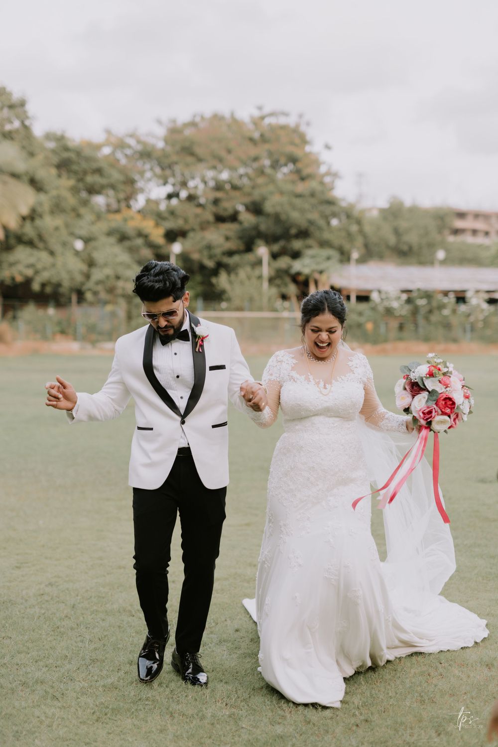 Newlyweds in white attire dancing joyfully across an outdoor field.