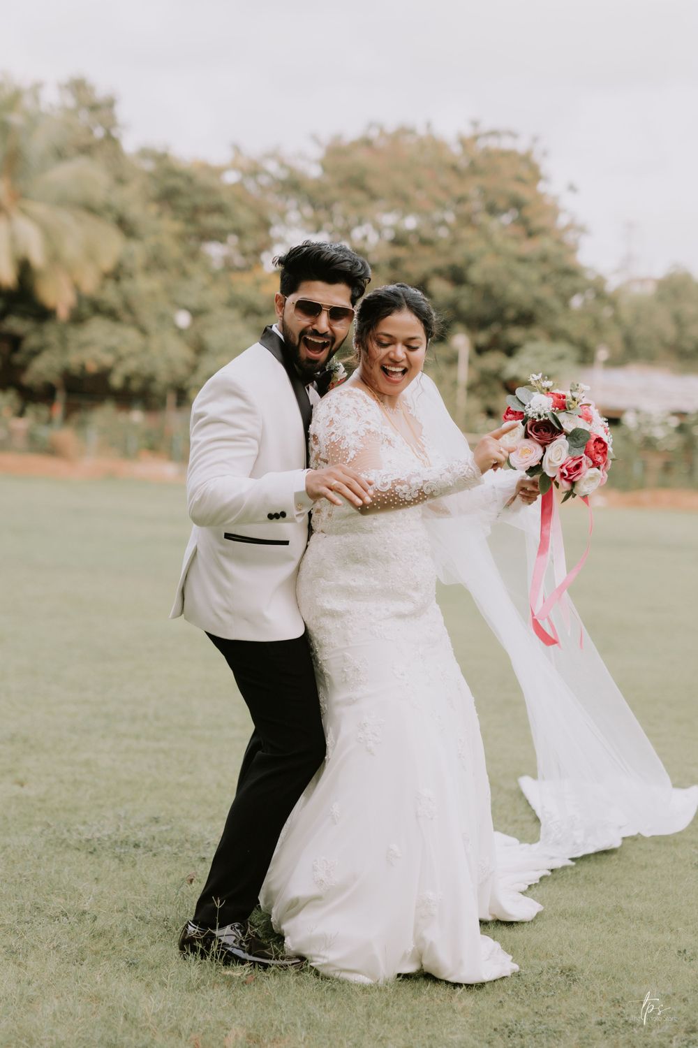 Wedding couple sharing laughter while dancing on green lawn with floral bouquet.