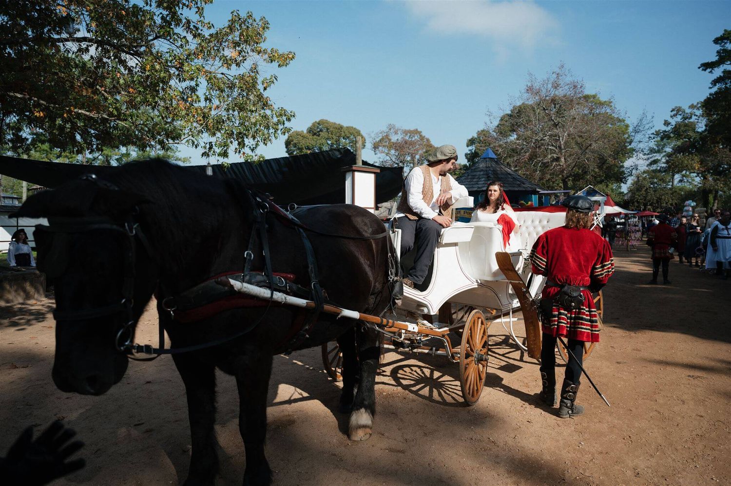 A black horse pulls an elegant white carriage along a dirt path at a renaissance festival.