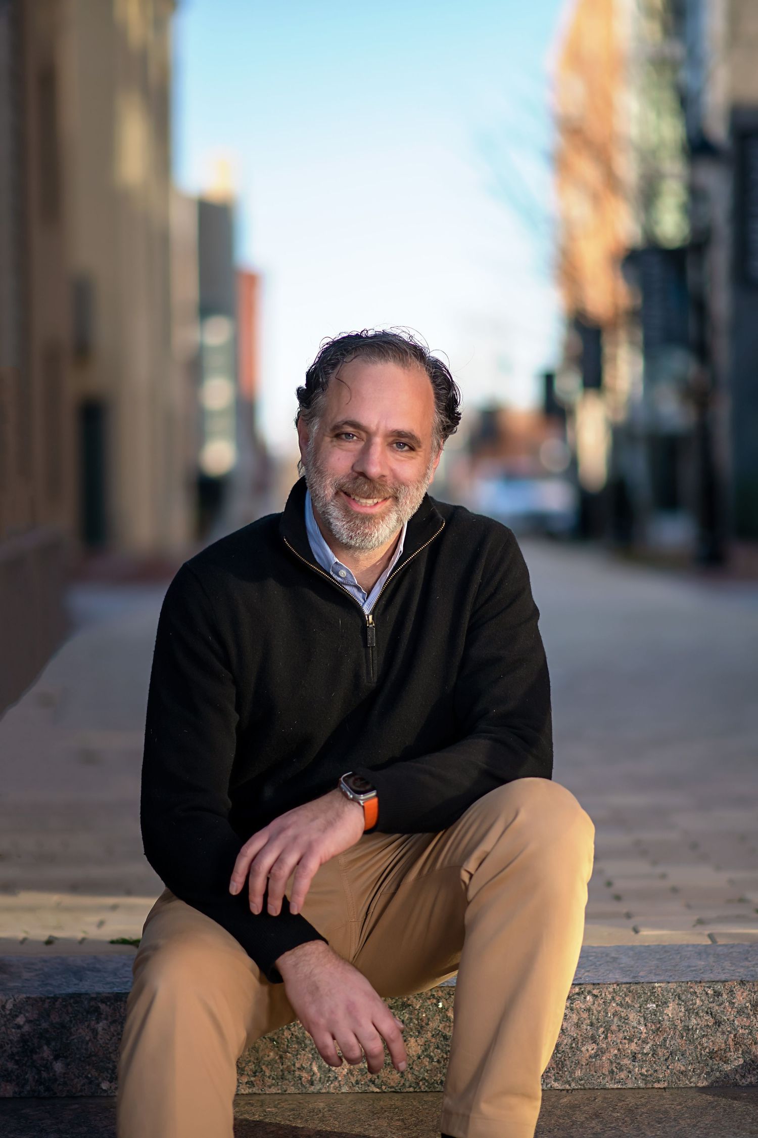 Casual outdoor portrait on cobblestone street with urban architecture in background.