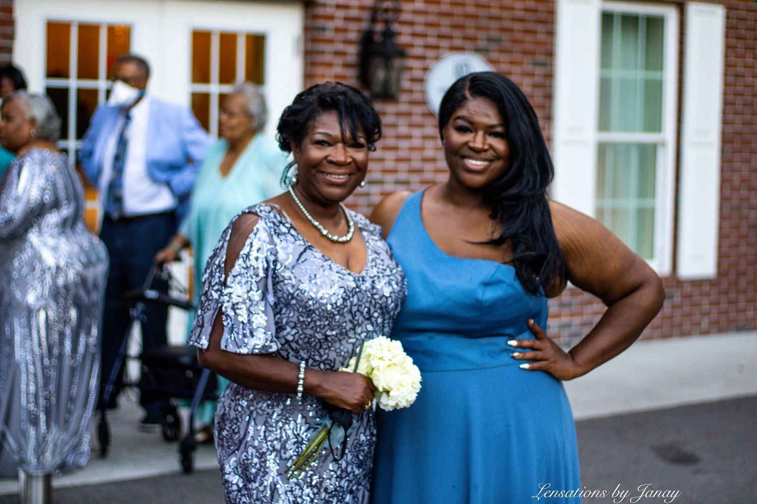 Two Black women smiling together at outdoor event, one in silver sequin dress and one in blue formal gown.