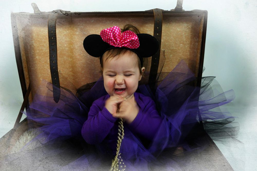 A baby wearing purple tulle and Minnie Mouse ears sits in an antique suitcase.