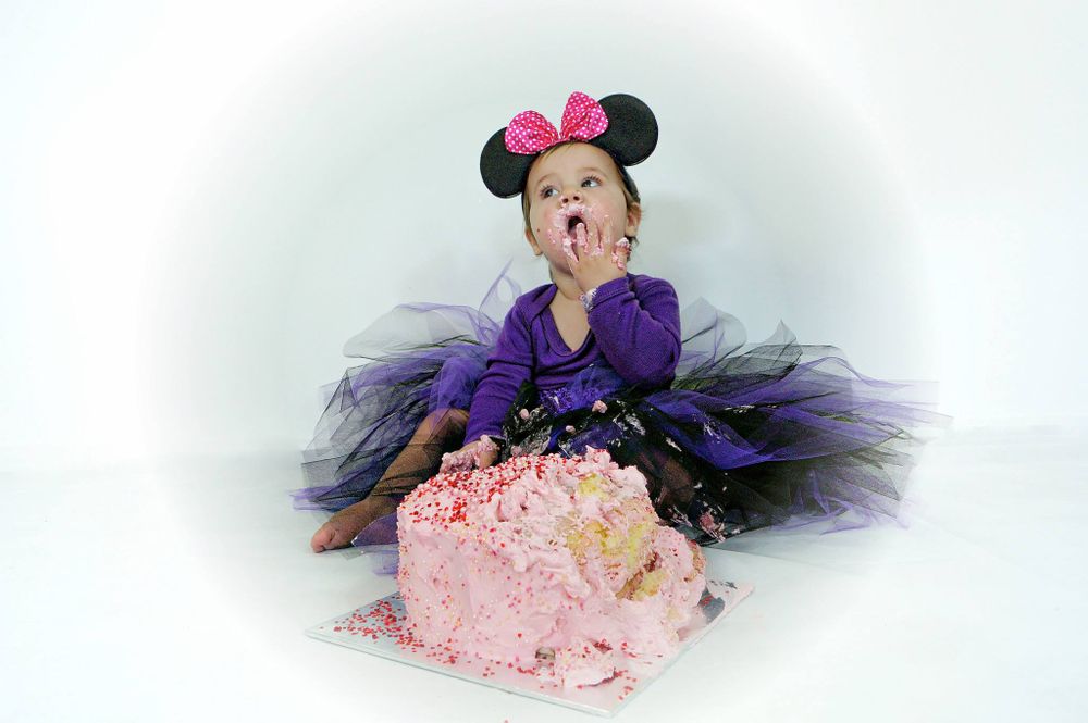 Baby in purple tutu and mouse ears sitting next to pink birthday cake.