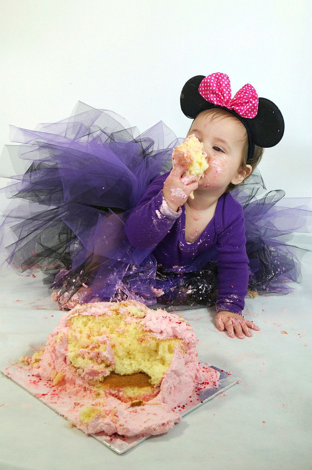 Baby in purple tutu and mouse ears enjoying smash cake photo session.