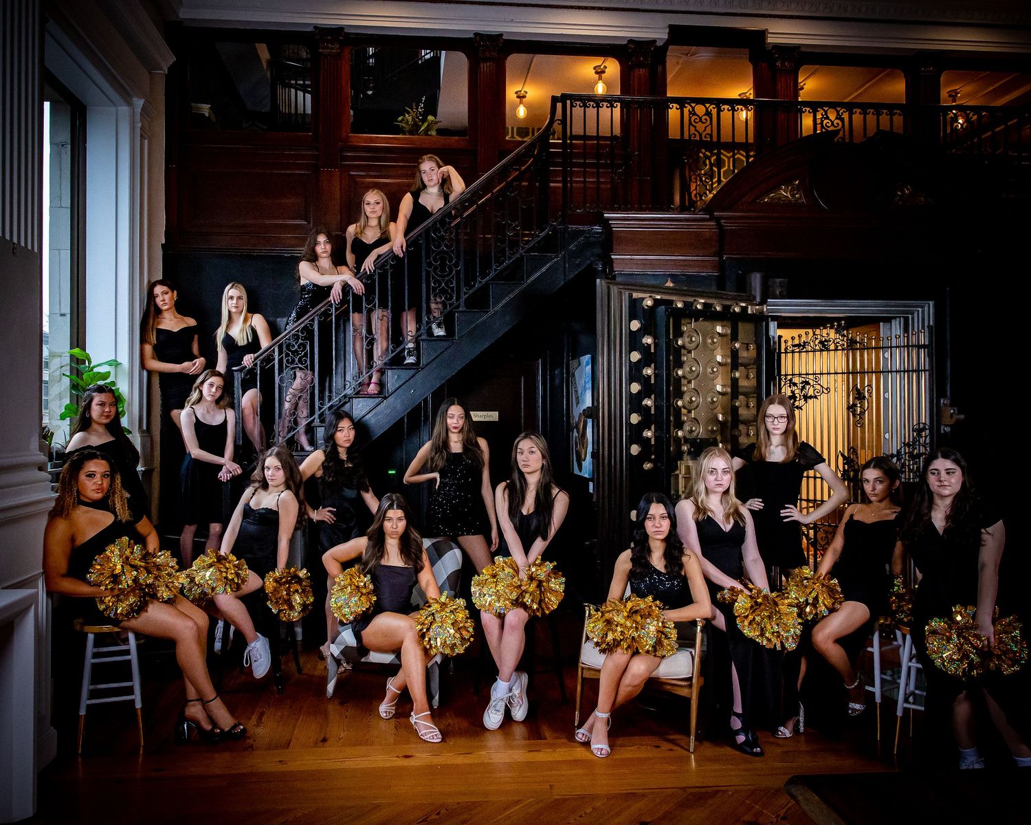 Dance team posing with gold pom poms on elegant staircase in dark venue.