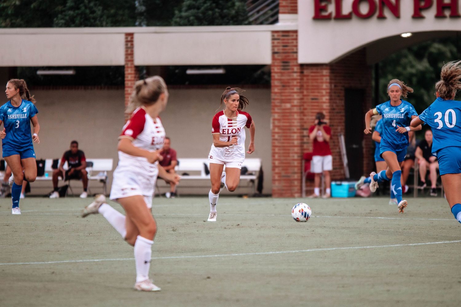 Soccer players in white and blue uniforms compete during a match at Elon Phoenix stadium.
