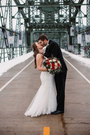 Romantic winter wedding kiss on bridge with red and white bouquet and industrial backdrop.