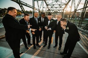 Groomsmen in black suits sharing laughs on bridge at sunset.