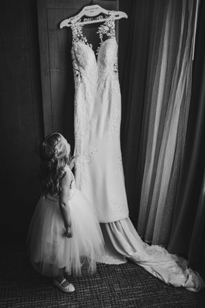 Wedding dress hanging with flower girl admiring it in black and white photo.