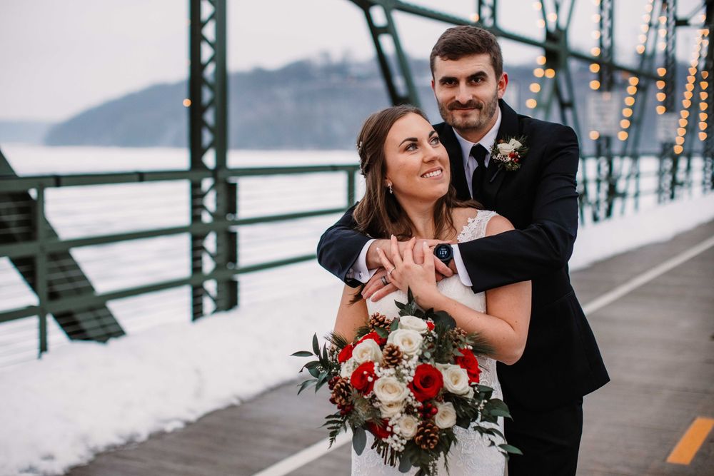 Newlyweds embrace on winter bridge with bride holding red and white bouquet.