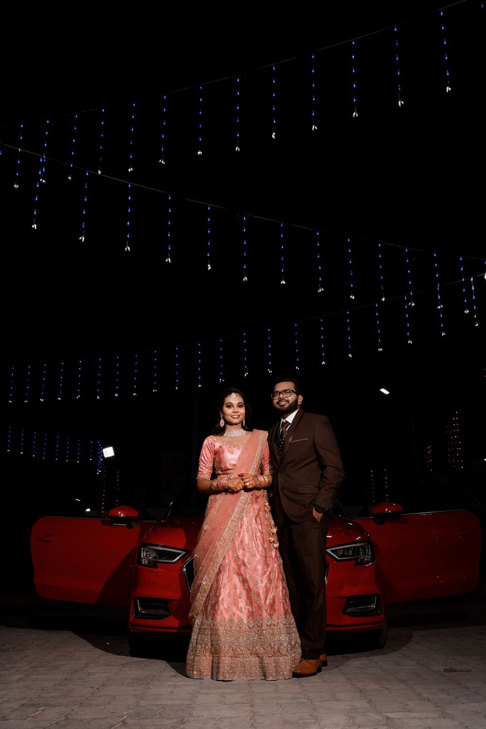 A couple in formal wedding attire pose in front of red cars under decorative string lights.