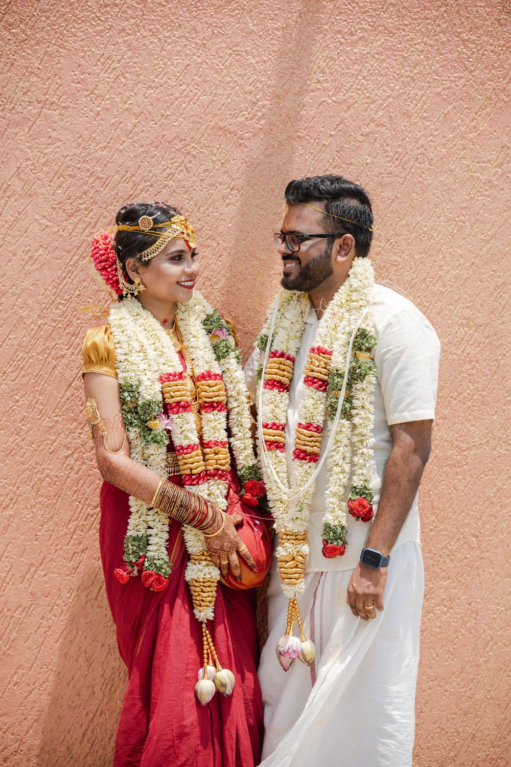 Couple in traditional South Indian wedding attire with flower garlands standing against coral wall.