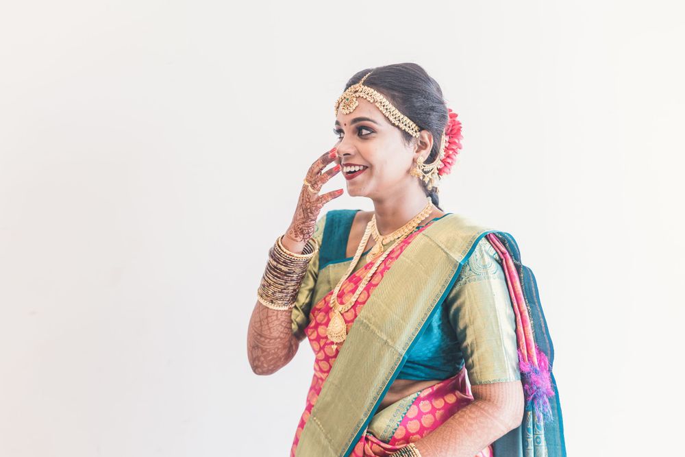 Woman in traditional silk saree with jewelry and flower in hair smiling against white background.