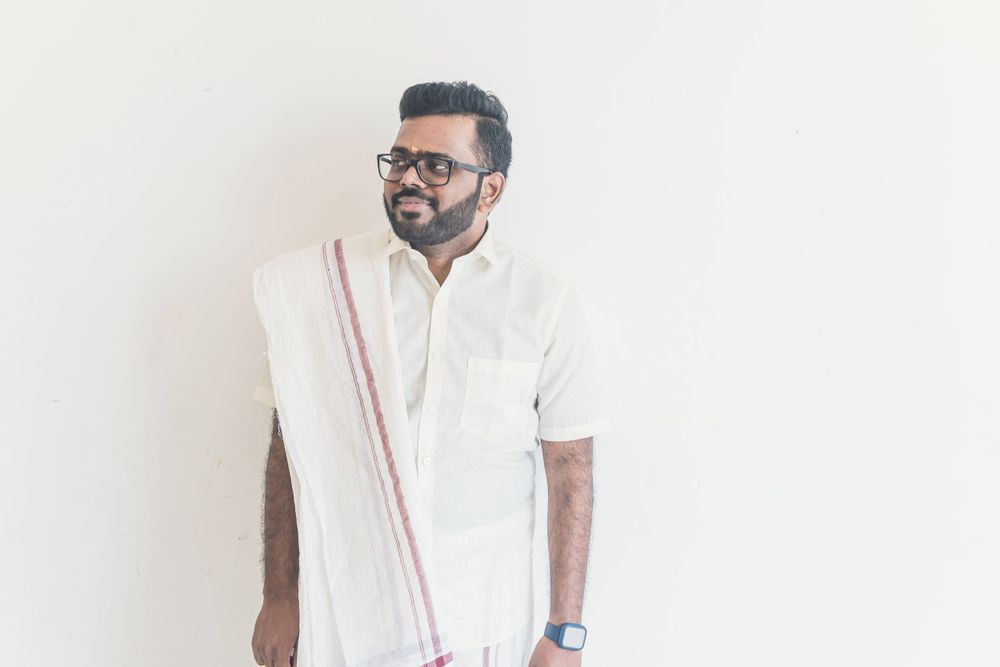 Man wearing white traditional Indian attire with glasses against plain wall.
