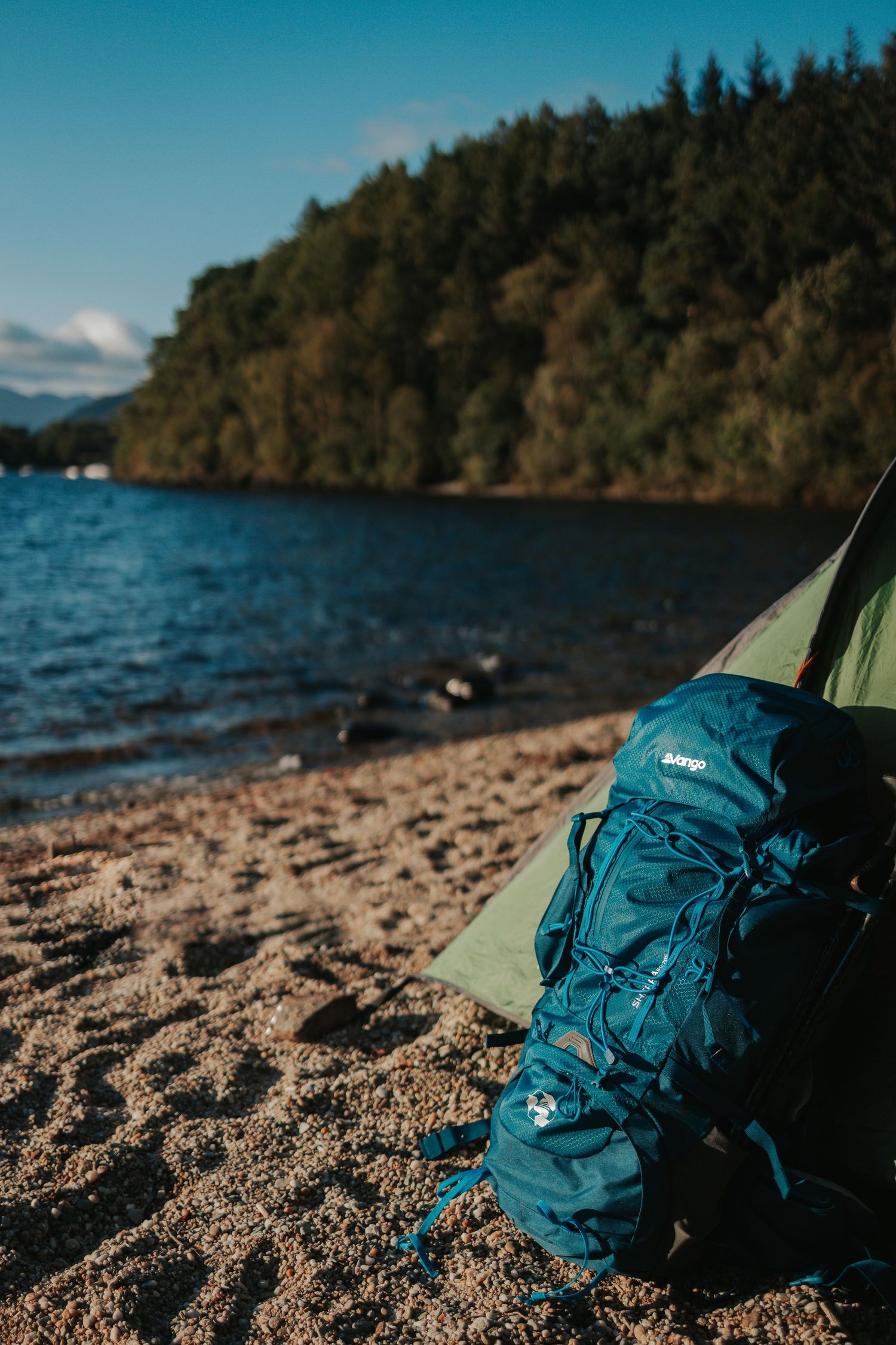 Blue hiking backpack rests against tent on sandy beach with forest and lake view.