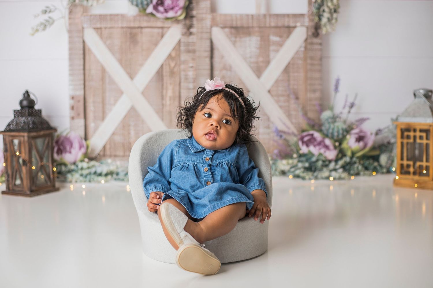 Baby in denim dress sits in gray container against rustic barn door and floral backdrop.