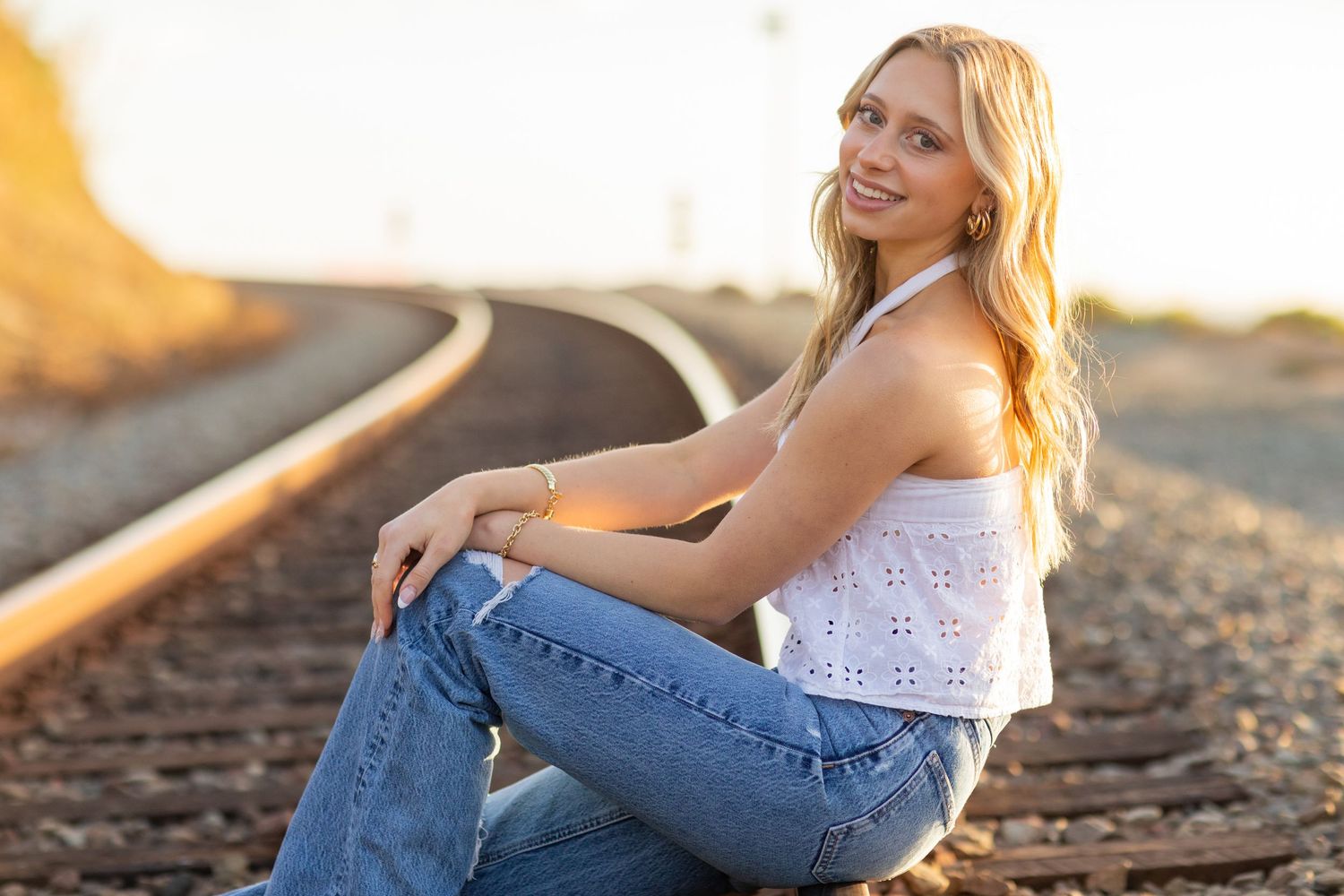 A high school senior in casual outfit sitting on railroad tracks during golden hour.