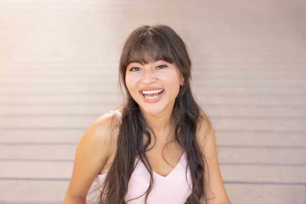 Woman with long dark hair and bangs smiling brightly against a neutral background.