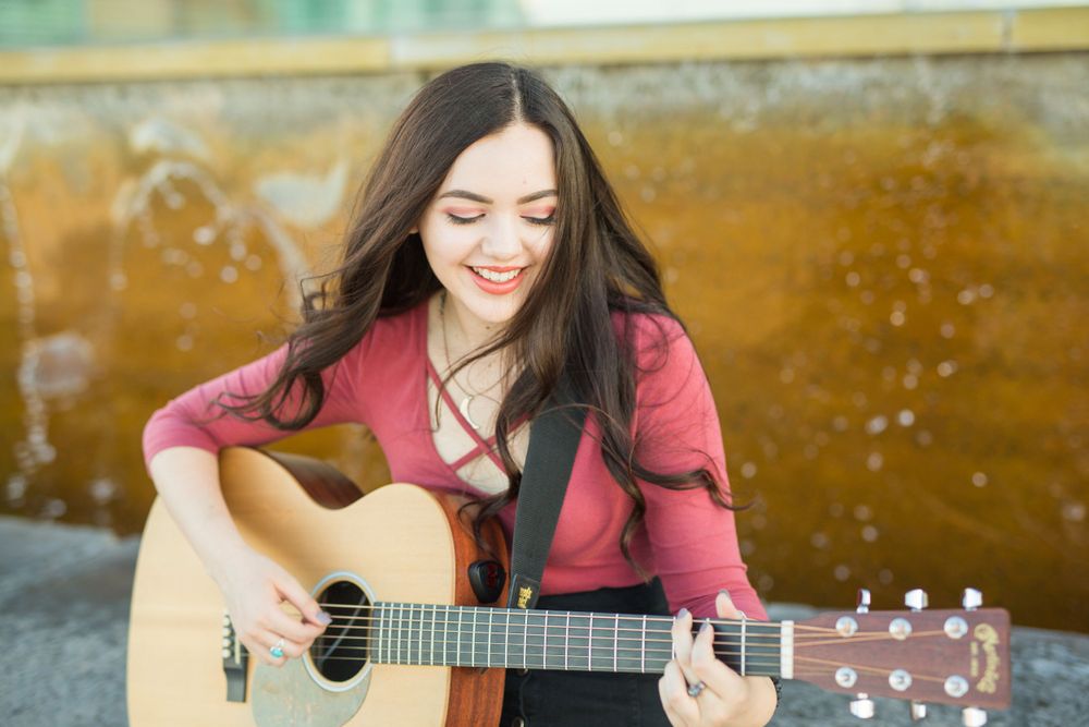 Person plays acoustic guitar while sitting outdoors with joyful expression.