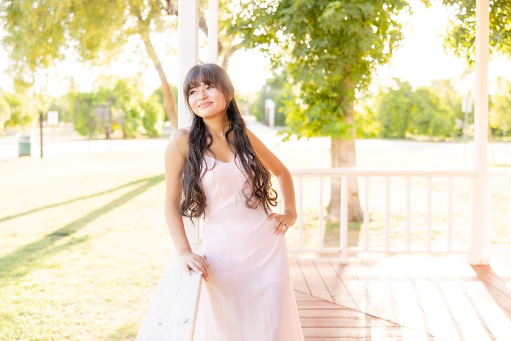 Woman in pink dress posing outdoors in warm sunlight beneath trees.