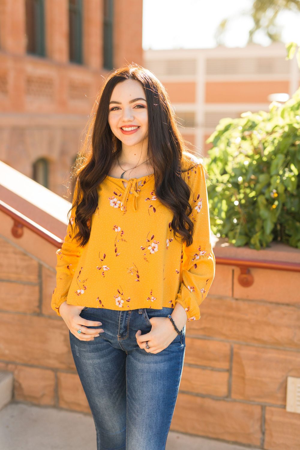 Person in yellow floral top and jeans poses in front of brick building.