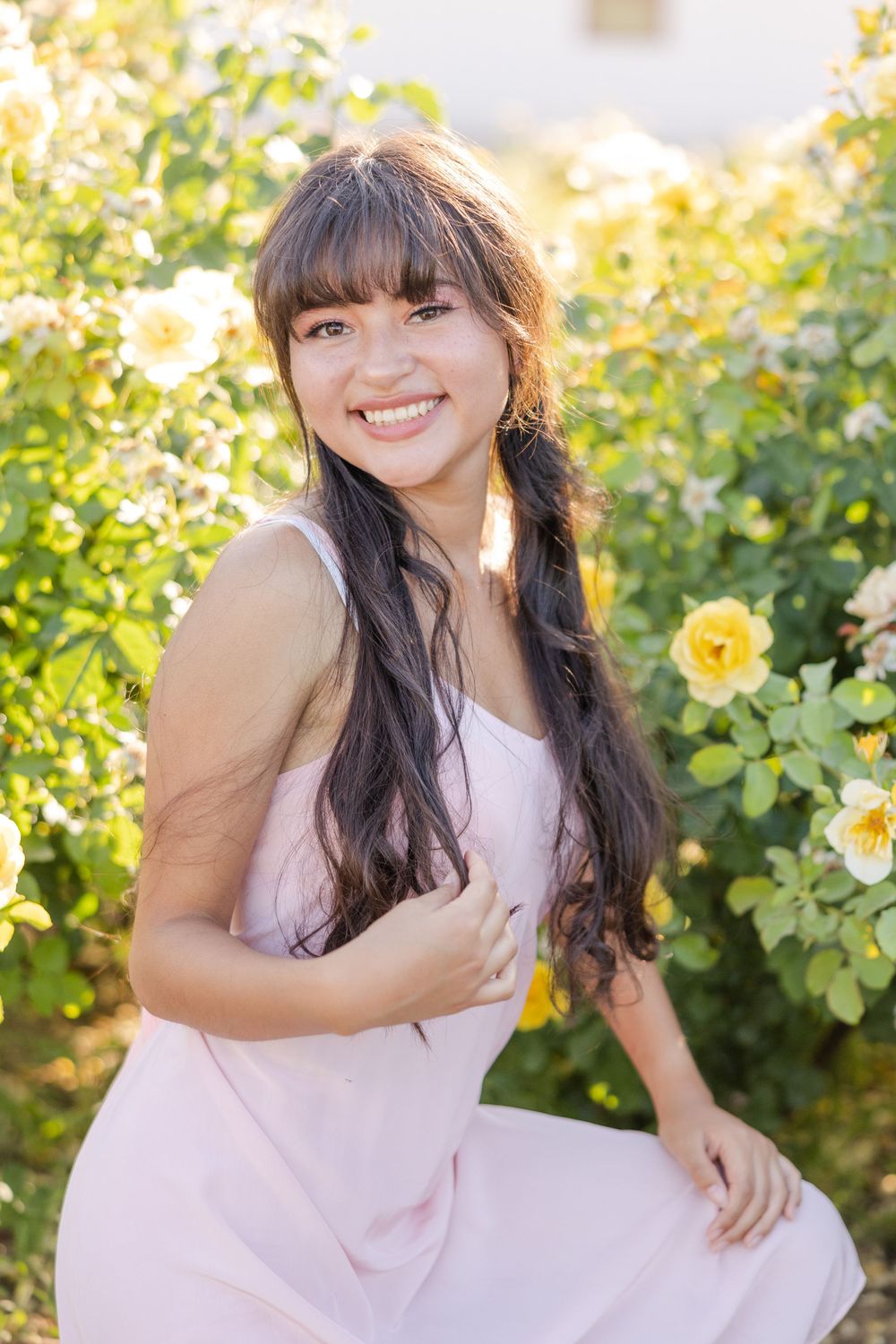 Woman in pink dress smiling among blooming yellow roses in garden setting.