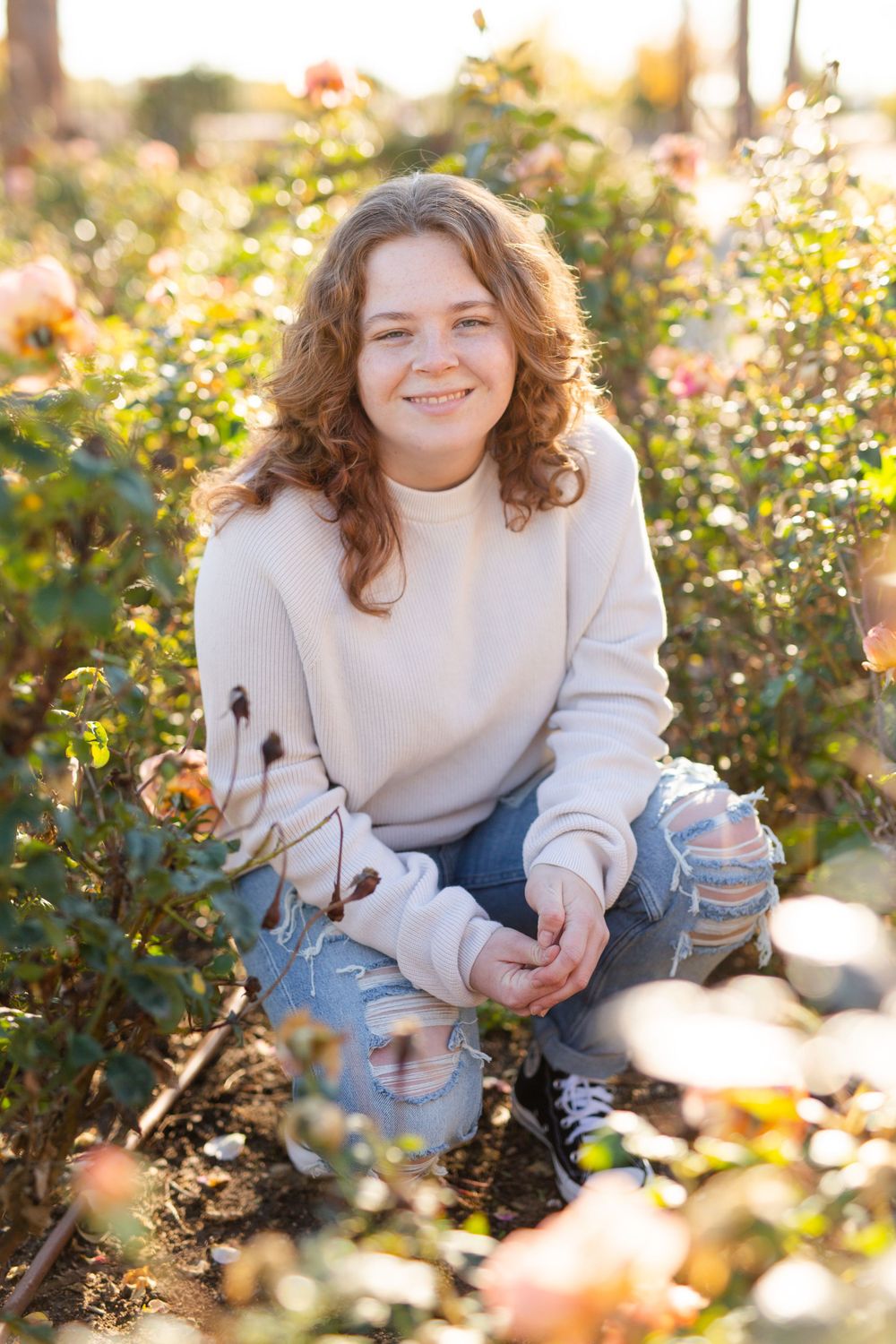 Person in cream sweater and ripped jeans poses in autumn flower garden.