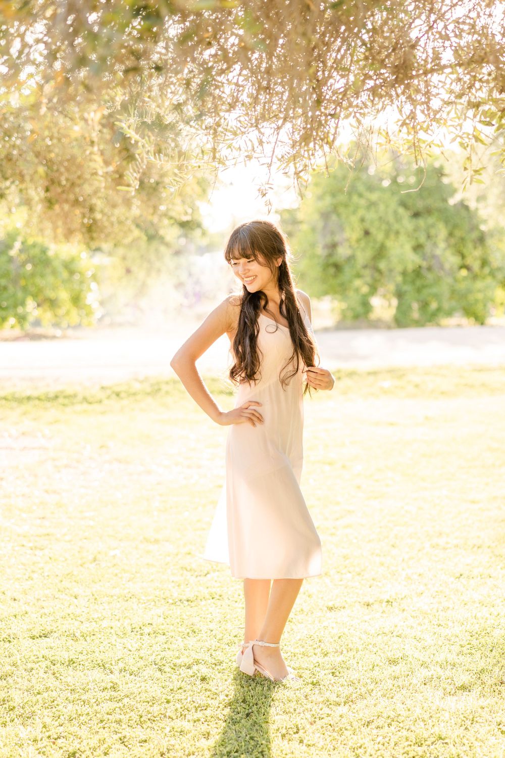 Woman in light dress posing in a sunlit garden with golden light filtering through trees.
