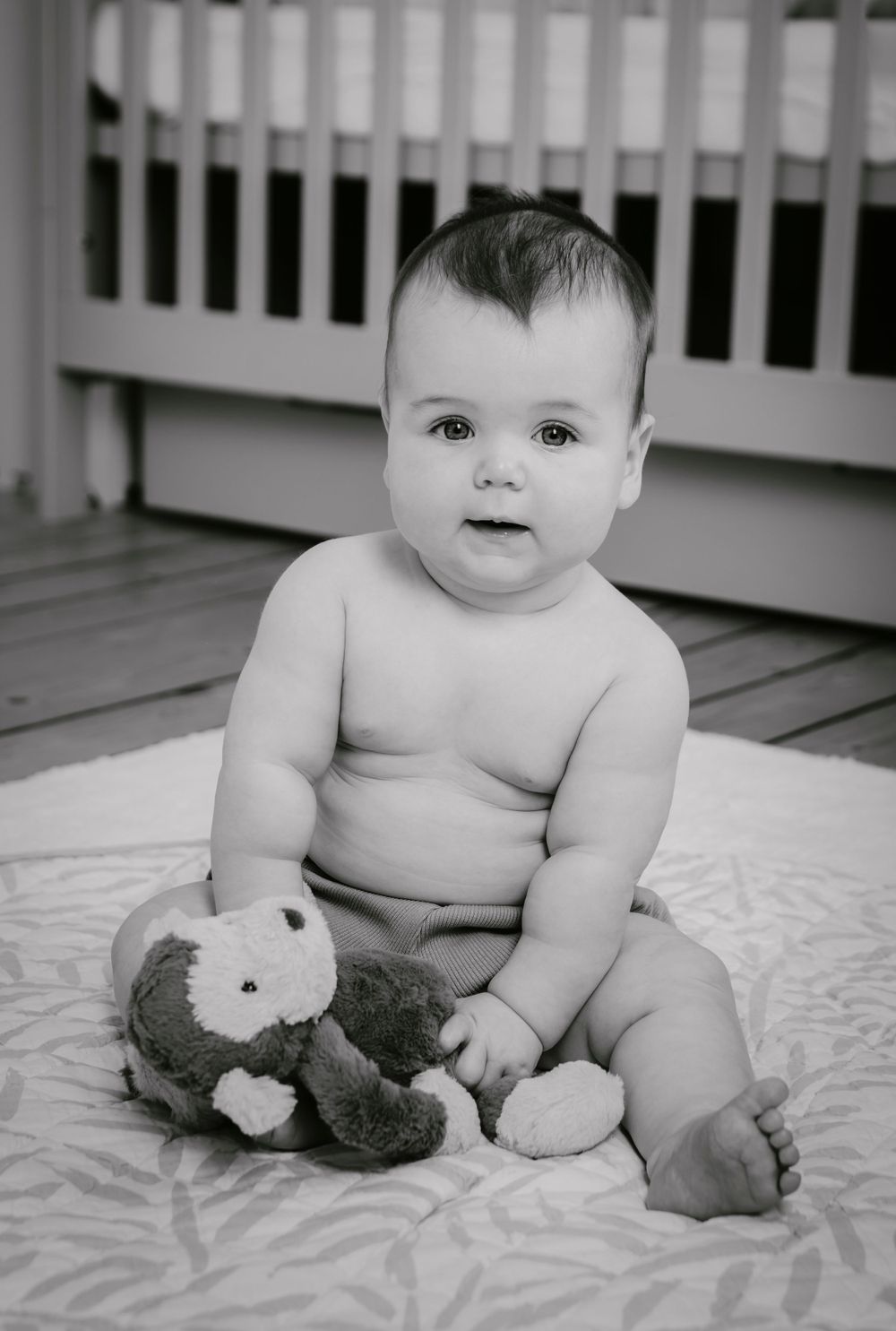 Black and white photo of infant sitting on blanket with plush toy by white crib.