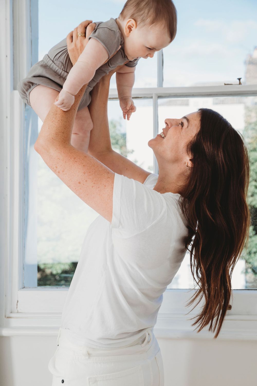 Parent in white shirt lifts baby in gray romper by bright window.