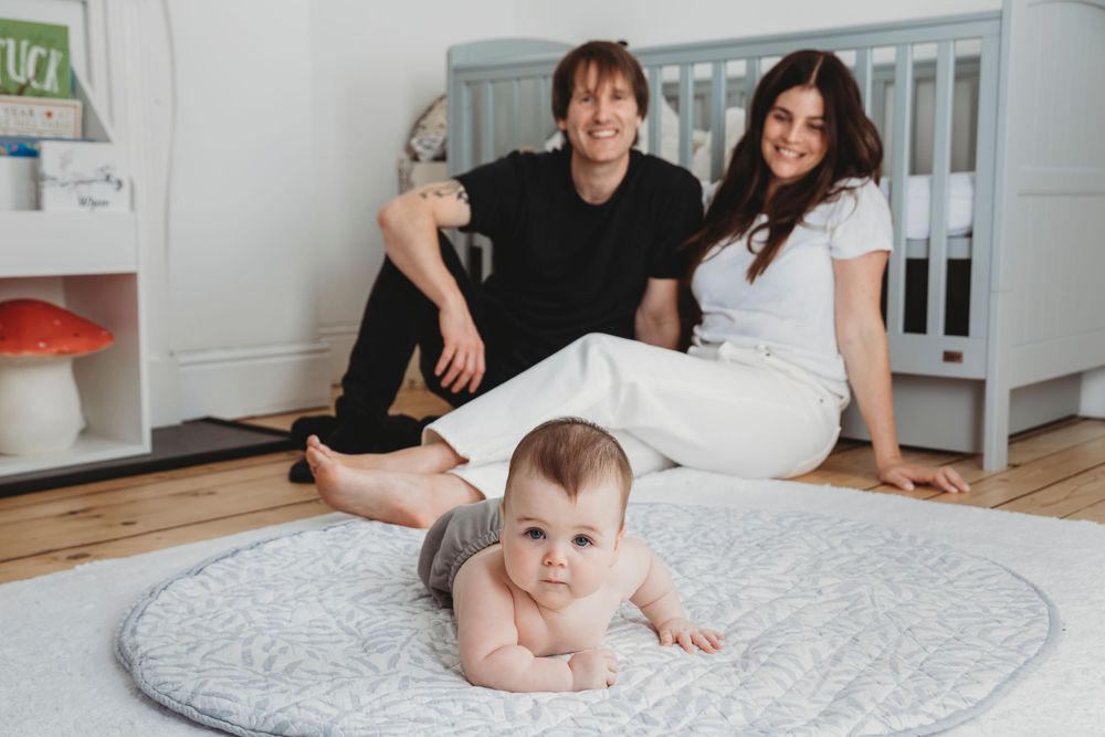 Baby lies on gray blanket on wooden floor while family sits on floor near white crib in nursery.