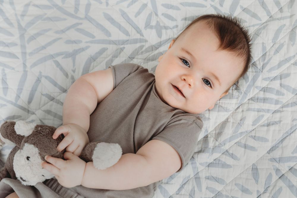 Baby lying on patterned blanket wearing grey onesie while holding plush toy.