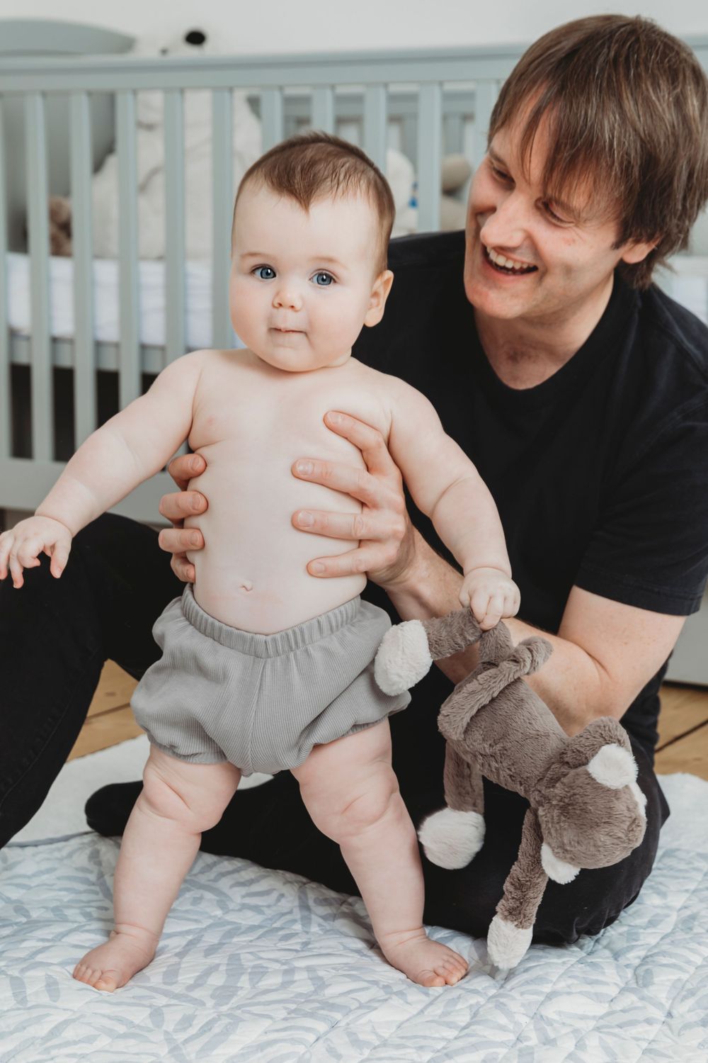 Baby in grey shorts standing with support while holding plush toy.