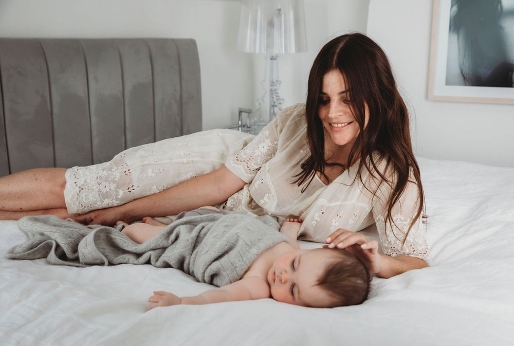 Parent and sleeping baby share tender moment while relaxing on gray bedding.