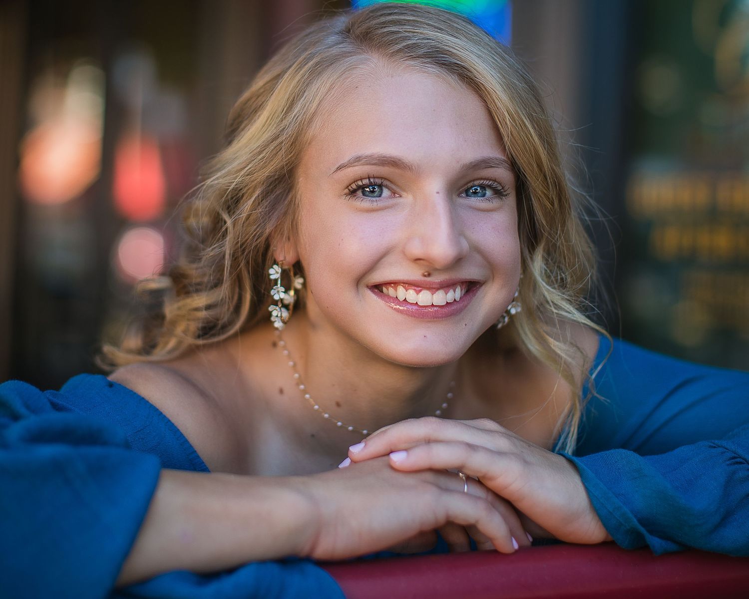 Portrait of young woman with blonde hair and dangling earrings smiling against blurred background.