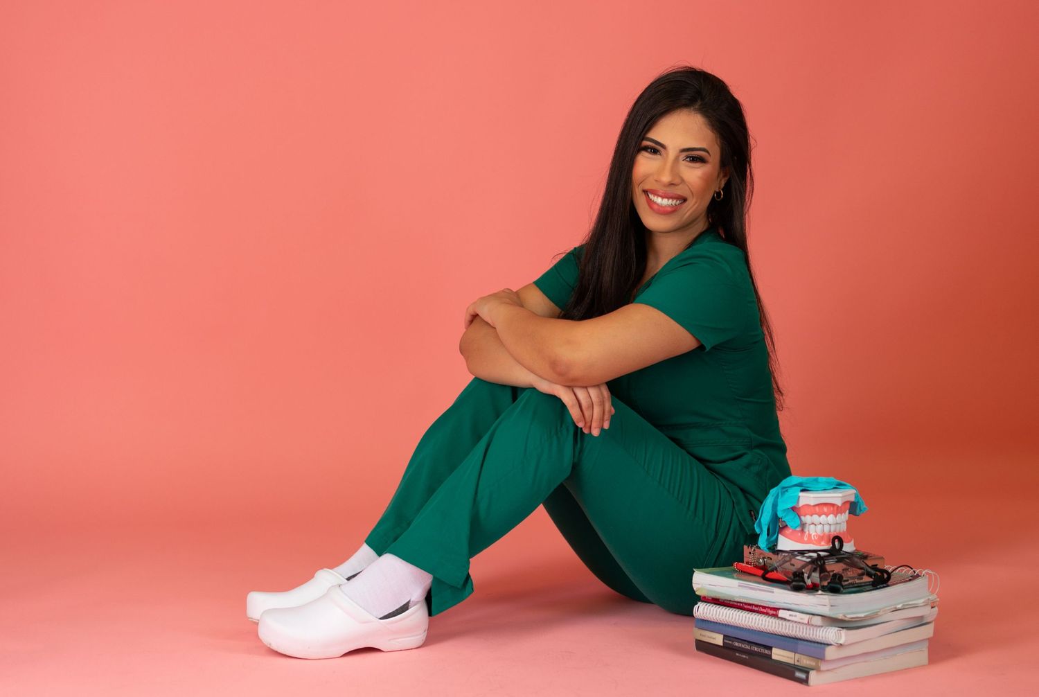Medical professional in green scrubs sits with books against coral background.