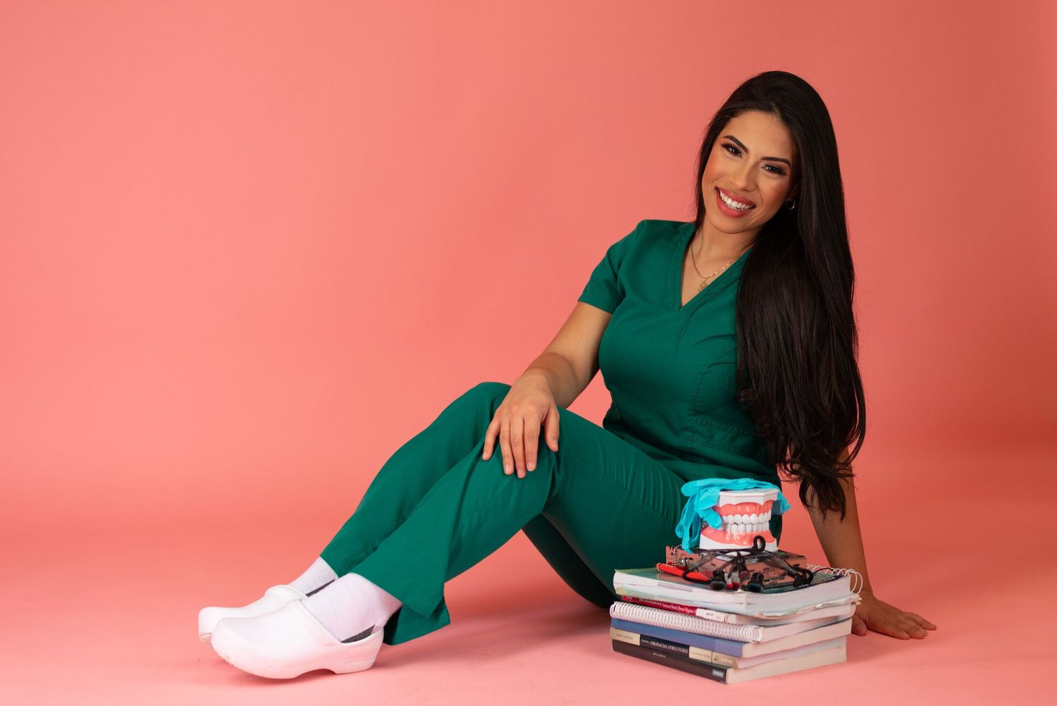 Healthcare worker in green uniform poses with medical textbooks on pink background.