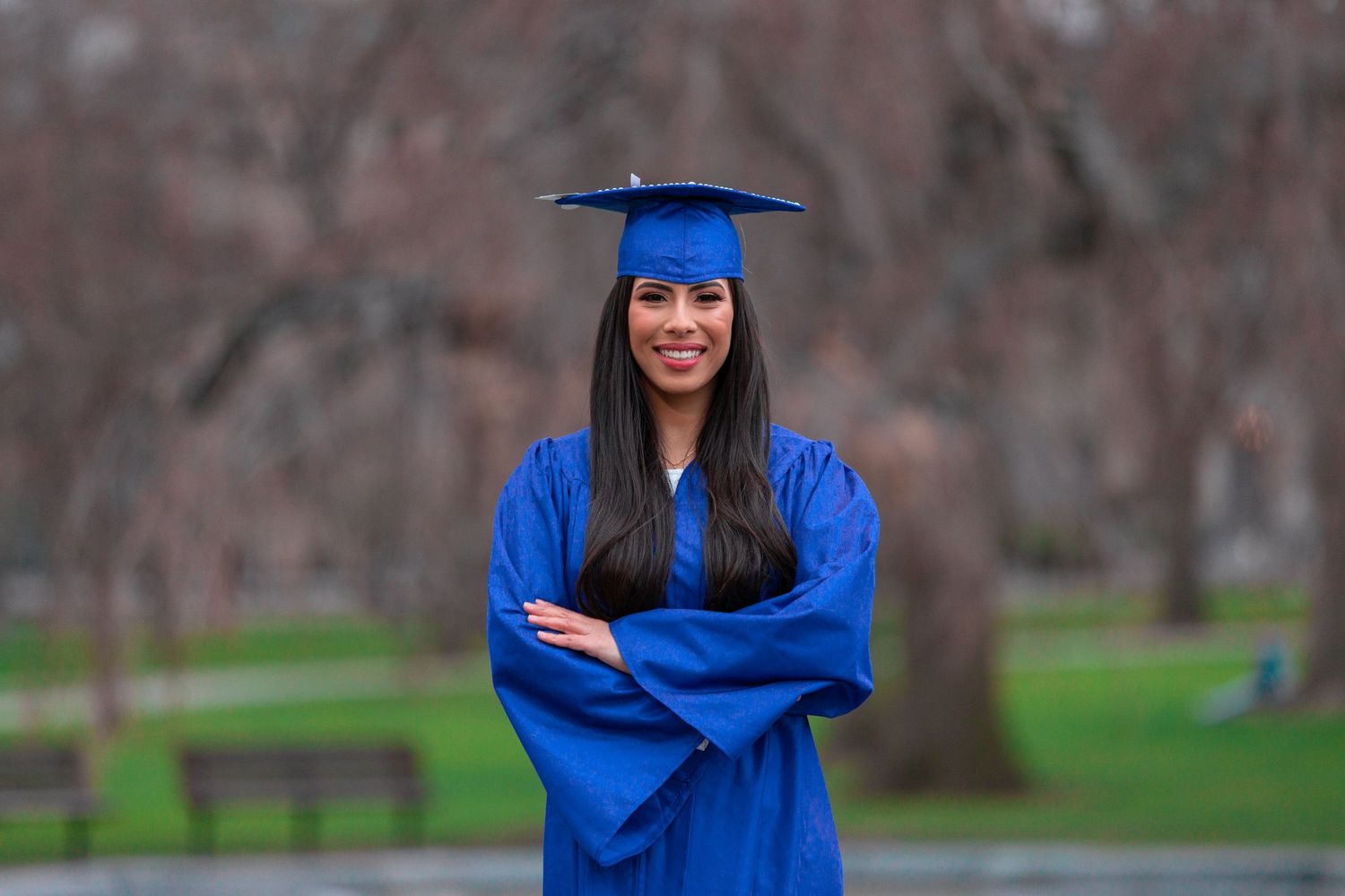 Graduate in blue cap and gown poses confidently with arms crossed outdoors.