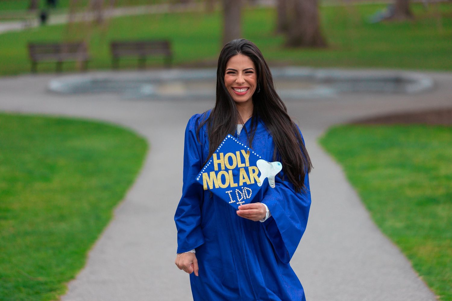 Dental graduate in blue regalia holds Holy Molar graduation cap decoration on campus path.