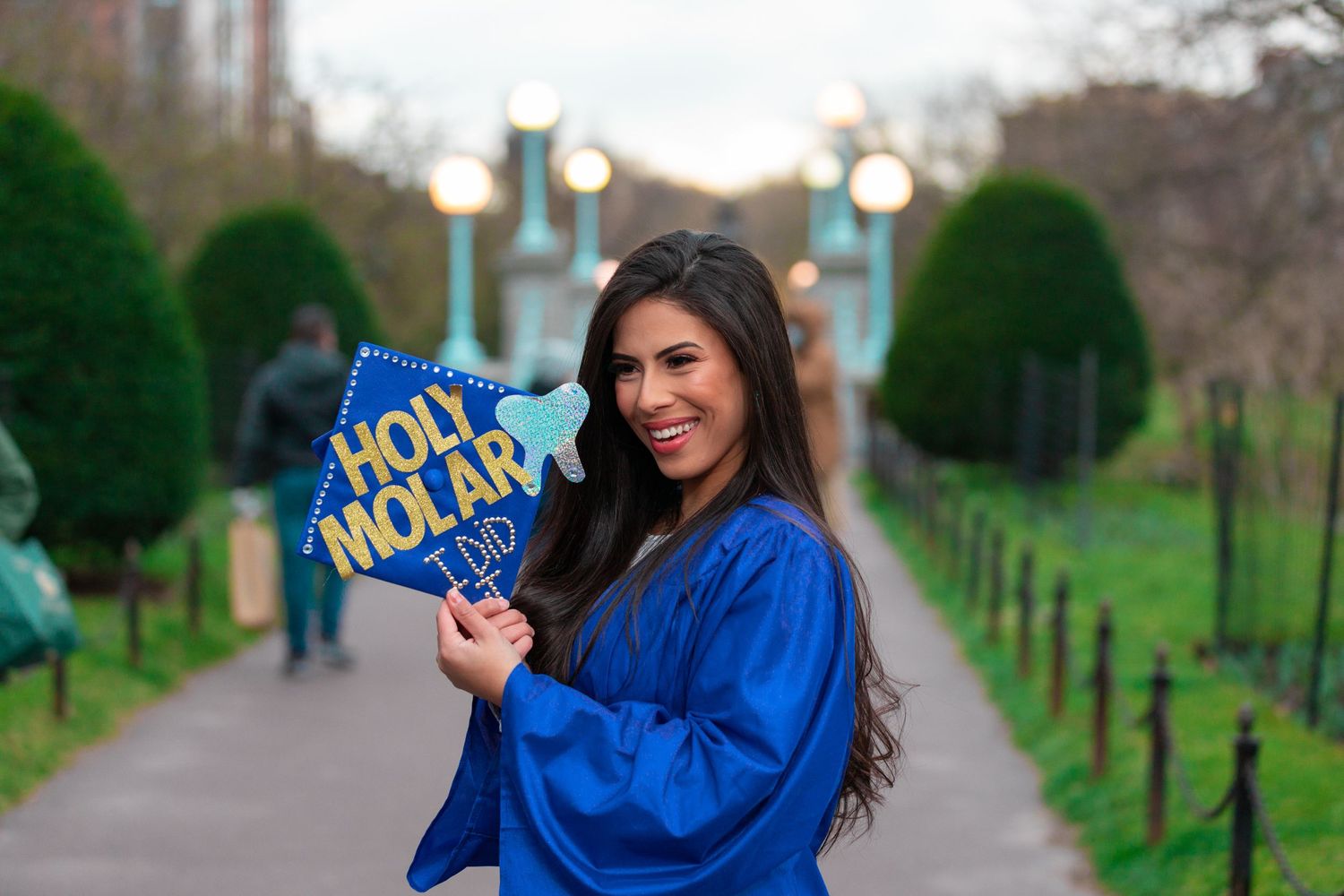 Graduate in blue commencement robe holds Holy Molar sign in garden setting.